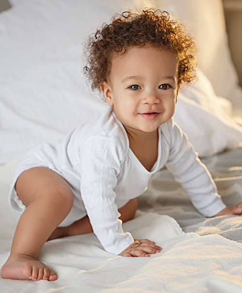 A smiling baby with curly hair wearing a white onesie, crawling on a bed with soft white linens