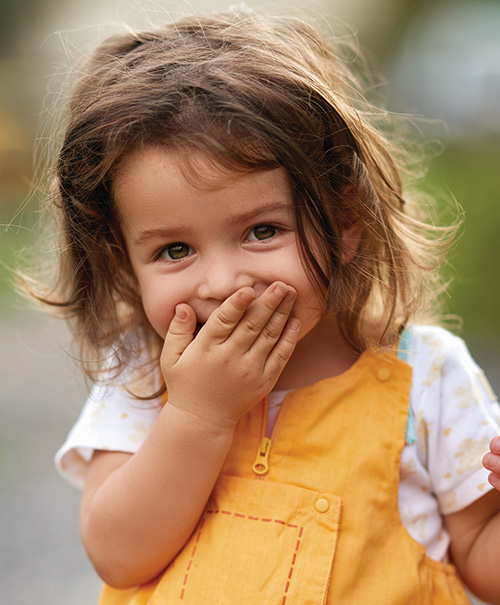A cheerful toddler with wavy hair, covering their mouth with one hand while smiling outdoors